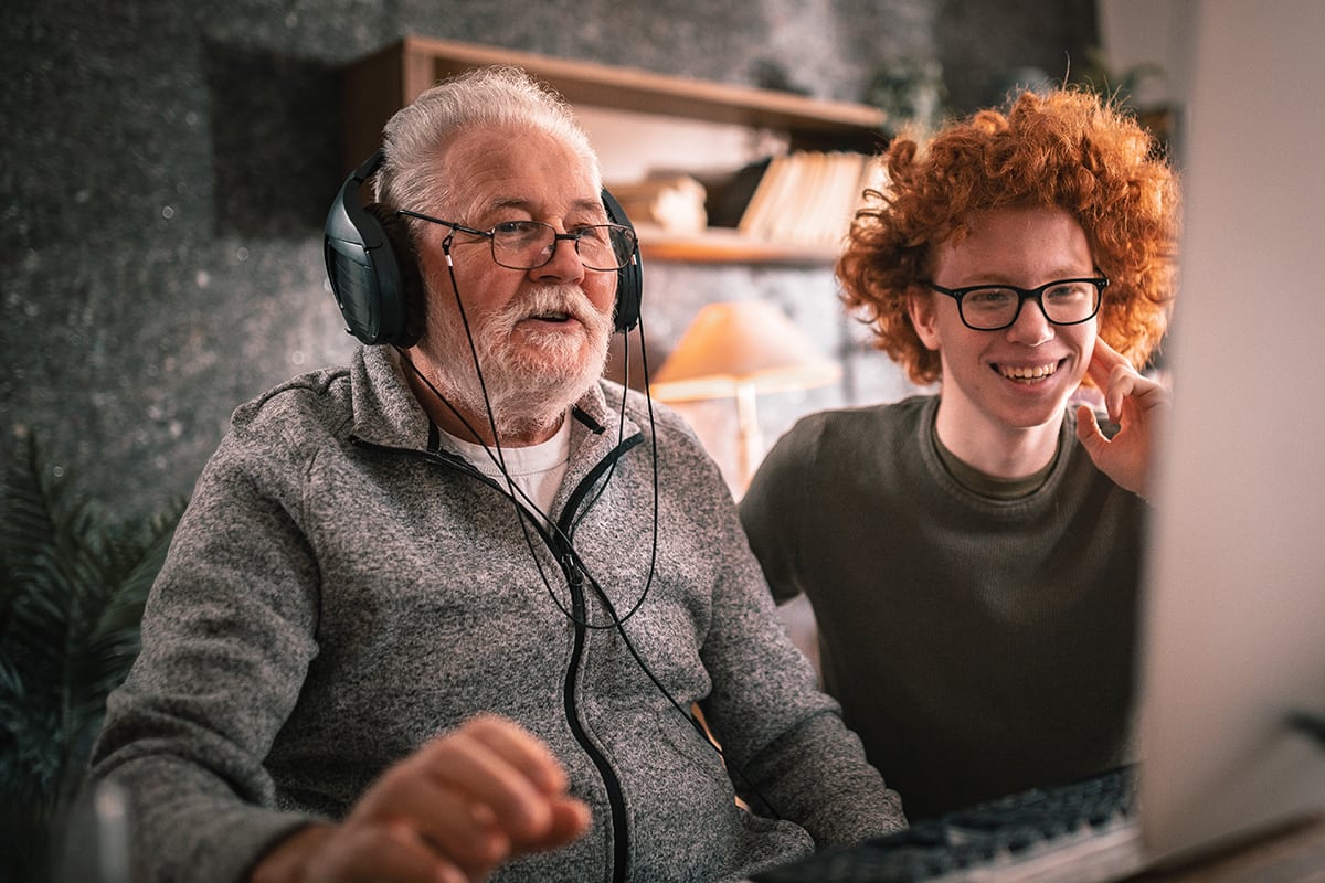 Young teen looks on as older man plays on computer with headphones